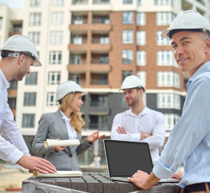 Smiling contented middle-aged engineer posing for the camera with his colleagues on the background
